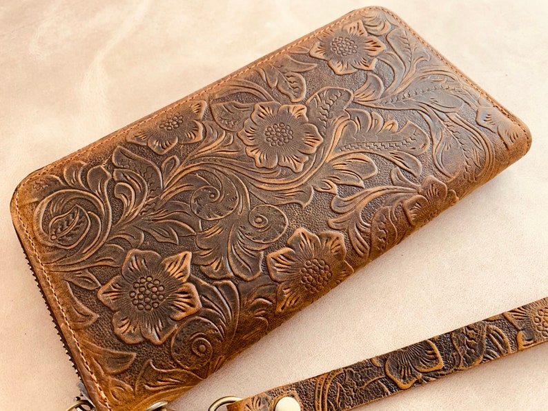 May include: A brown leather wallet with a floral embossed design. The wallet has a zipper closure and a wrist strap.