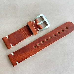 May include: A brown leather watch strap with a silver buckle. The strap has white stitching and multiple holes for adjustment. The leather has a textured appearance.