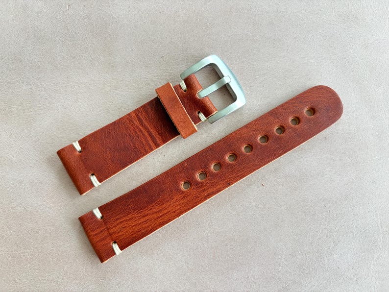 May include: A brown leather watch strap with a silver buckle. The strap has white stitching and multiple holes for adjustment. The leather has a textured appearance.