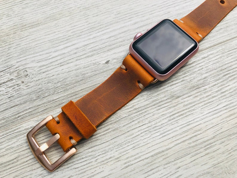 May include: A brown leather watch band with a rose gold buckle and a rose gold Apple Watch.