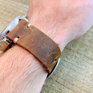 May include: A brown leather watch strap with a stainless steel watch face. The strap is distressed with visible grain and stitching. The watch is on a wrist.