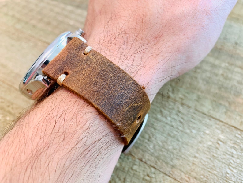 May include: A brown leather watch strap with a stainless steel watch face. The strap is distressed with visible grain and stitching. The watch is on a wrist.