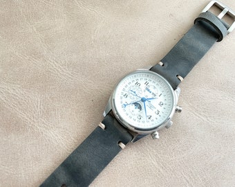 May include: A silver chronograph wristwatch with a white face and black leather band. The watch has a moon phase indicator, a date window, and a 24-hour subdial. The watch is on a light brown surface.