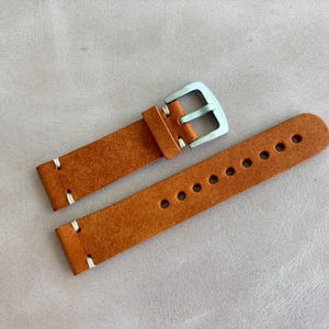May include: A pair of new, brown leather watch straps with white stitching and a silver-colored buckle. The strap on the right has multiple holes for adjustment. The watch straps are displayed on a light gray surface.