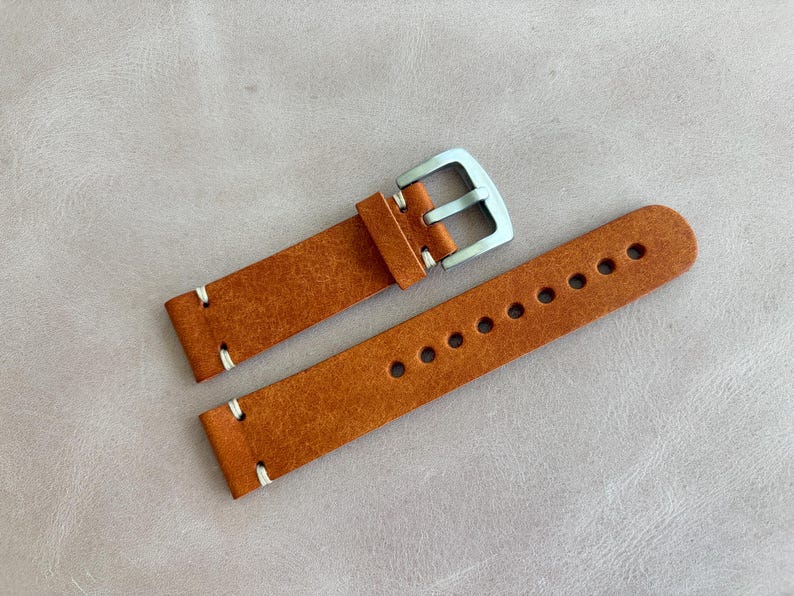 May include: A pair of new, brown leather watch straps with white stitching and a silver-colored buckle. The strap on the right has multiple holes for adjustment. The watch straps are displayed on a light gray surface.