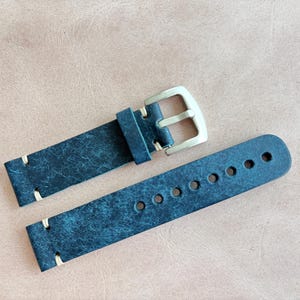 May include: A distressed navy blue leather watch strap with a silver-colored buckle. The strap has cream-colored stitching and multiple adjustment holes. The watch band is displayed on a light brown surface.