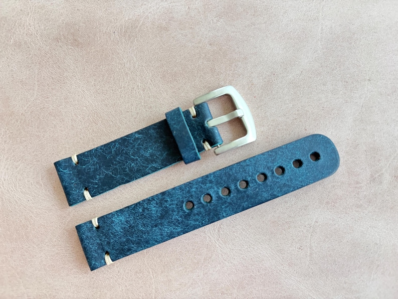 May include: A distressed navy blue leather watch strap with a silver-colored buckle. The strap has cream-colored stitching and multiple adjustment holes. The watch band is displayed on a light brown surface.