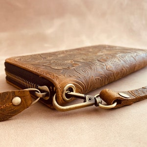 May include: A brown leather wallet with a floral embossed design and a zipper closure. The wallet has a detachable strap with a metal clasp.