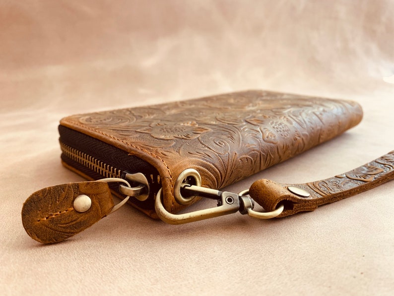 May include: A brown leather wallet with a floral embossed design and a zipper closure. The wallet has a detachable strap with a metal clasp.