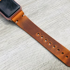 May include: A brown leather watch band with multiple holes for adjusting the fit. The band is attached to a rose gold Apple Watch.