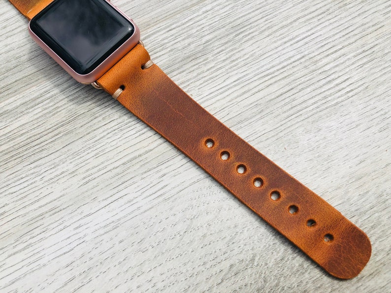 May include: A brown leather watch band with multiple holes for adjusting the fit. The band is attached to a rose gold Apple Watch.