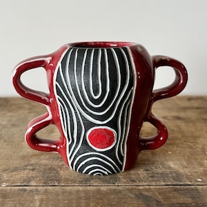 Może przedstawiać: Czerwony ceramiczny wazon z dwoma uchwytami i abstrakcyjnym wzorem w kolorze czarnym i białym. Wzór zawiera czerwone koło w środku.