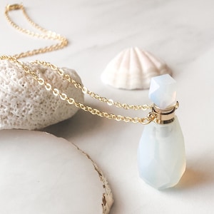 May include: A gold chain necklace with a white, faceted, glass bottle pendant. The bottle has a gold stopper and is shaped like a teardrop. The necklace is laying on a white surface with a seashell and a piece of coral.