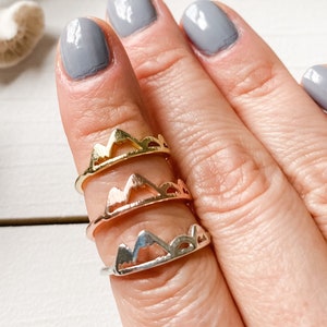 Puede incluir: Tres anillos de oro con silueta de montaña apilados en un dedo. Los anillos son de diferentes tonos de oro, siendo el anillo superior el más claro y el anillo inferior el más oscuro.