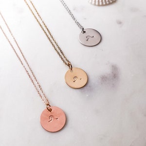 May include: Three delicate necklaces with a wave design pendant. The necklaces are silver, gold, and rose gold. The necklaces are displayed on a white surface with seashells.