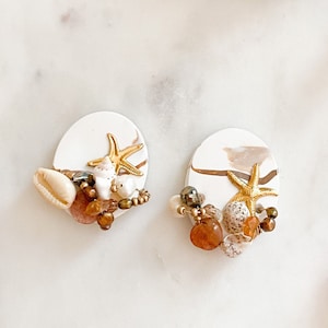 Puede incluir: Un par de pendientes de arcilla blanca con un diseño de concha y estrella de mar. Los pendientes están decorados con cuentas marrones y doradas y tienen un efecto marmoleado.