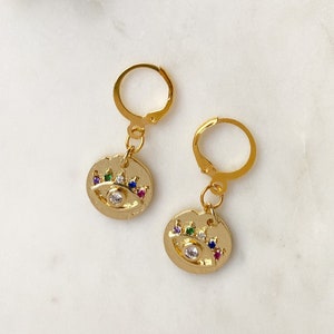 May include: A pair of gold hoop earrings with a small charm hanging from each hoop. The charms are round and feature a gold eye with a white centre and colourful gemstones around the eye.
