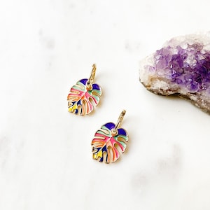 May include: A pair of gold-tone earrings featuring a colourful enamel monstera leaf design. The leaves are outlined in gold and have a variety of colours, including pink, blue, green, and yellow.