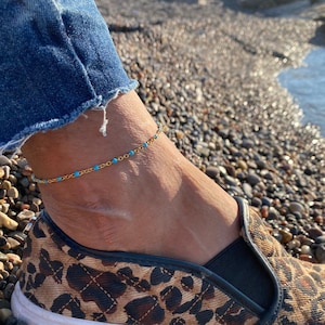 Puede incluir: Un tobillo de cadena dorada con pequeñas cuentas azules. El tobillo se lleva en el tobillo de una persona, con un zapato de estampado de leopardo en el pie.