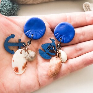 May include: A pair of blue and brown earrings with a nautical theme. Each earring features a blue circle with a textured surface, a wooden anchor, a wooden sailboat, and a variety of seashells.