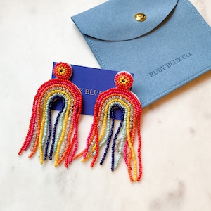 May include: Rainbow-shaped beaded earrings in red, orange, yellow, and blue. The earrings feature a fringe design and are presented with a blue pouch. The pouch has the text "RUBY BLUE CO."