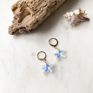 Puede incluir: Un par de pendientes de aro dorados con abalorios de estrella de mar de cristal iridiscente. Los pendientes están sobre una superficie blanca con un trozo de madera flotante y una concha.