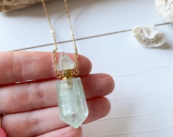 May include: A gold chain necklace with a clear crystal pendant shaped like a bottle. The bottle has a gold stopper and is filled with a light green liquid.