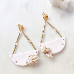 May include: A pair of white ceramic earrings with gold accents and a seashell charm. The earrings are shaped like a crescent moon and have a gold bar hanging from the top. The seashell charm is attached to the bottom of the bar.