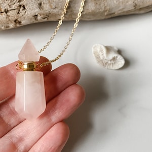 Op de afbeelding: Een ketting met een hanger van rozenkwartskristal, een gouden ketting en een gouden dop. De hanger heeft de vorm van een fles en heeft een puntige bovenkant.