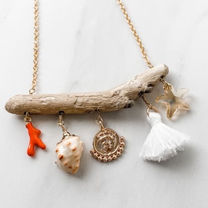 Puede incluir: Un collar de cadena dorada con un colgante de madera flotante. El colgante tiene un borla blanca, una concha, un coral y un amuleto de moneda de oro.