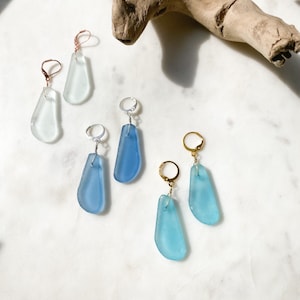 May include: Three pairs of earrings with different coloured glass teardrop shapes. The earrings are on a white surface with a piece of driftwood in the background. The earrings are made with silver, gold, and rose gold.