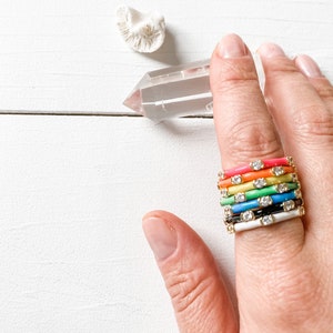 May include: A stack of colourful enamel rings with small clear stones on a white background.