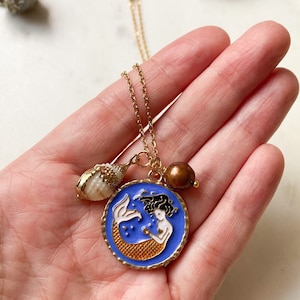May include: Gold chain necklace with a blue and gold mermaid charm, a small seashell charm, and a brown pearl charm.