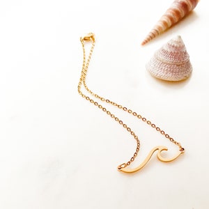May include: A gold-colored necklace with a wave pendant, displayed on a white surface. Two seashells in shades of brown and pink are in the background. The necklace chain is delicate.