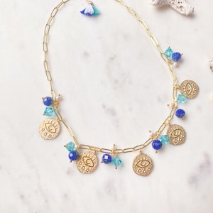 May include: Gold chain necklace with a blue and white tassel charm. The necklace features multiple gold charms with an evil eye design, blue and turquoise beads, and clear crystal beads.