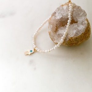 May include: A delicate pearl bracelet with a gold hamsa hand charm featuring a blue evil eye. The bracelet is displayed on a white marble surface with a geode.