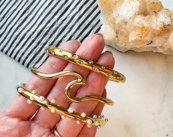 Può includere: Tre braccialetti d'oro impilati su una mano. Il braccialetto superiore ha piccole pietre trasparenti, il braccialetto centrale ha la forma di un'onda e il braccialetto inferiore ha piccole perle bianche.
