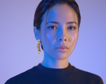 May include: A woman with dark hair wearing a black mock neck top and gold leaf earrings. The woman is illuminated with blue light.