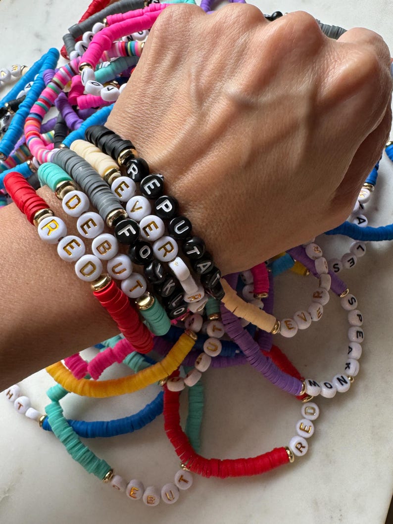May include: A stack of colorful beaded bracelets with letter beads spelling out words like "REPUTA", "EVER", "FOKLO", "DEBUT", "RED", "HT", "LES", "EVIE", "S", "M", "T", "D", "K", "MOND", and "MRED".