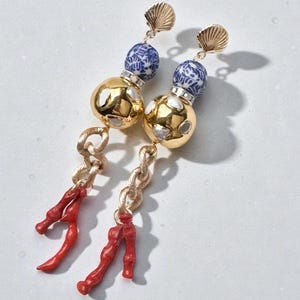 May include: A pair of dangle earrings featuring gold-colored metal, blue and white ceramic beads, and red coral accents. The earrings have a seashell-shaped top and a chain-like design.