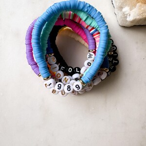 May include: A set of colorful beaded bracelets with letter and number charms. The bracelets are made of light blue, purple, pink, and green beads. The charms spell out the words "MIDNIGHT" and "FOLLOW" and include the numbers "1989".