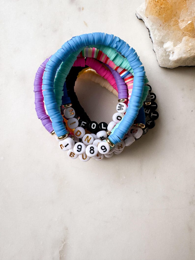 May include: A set of colorful beaded bracelets with letter and number charms. The bracelets are made of light blue, purple, pink, and green beads. The charms spell out the words "MIDNIGHT" and "FOLLOW" and include the numbers "1989".