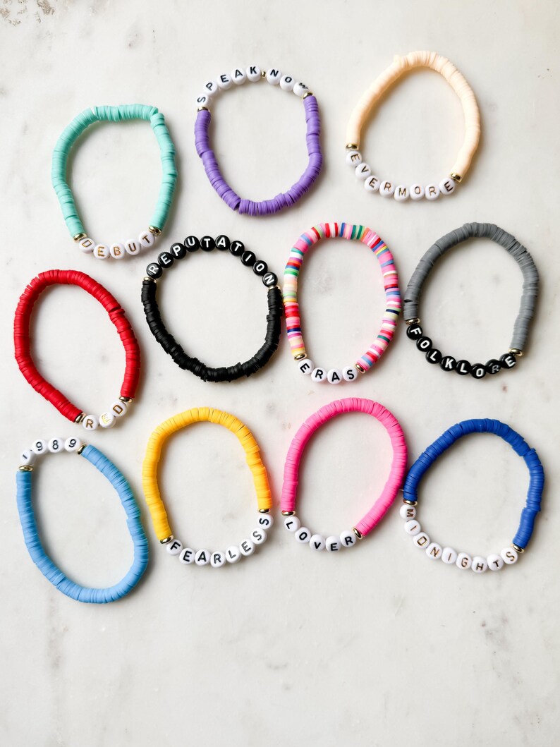 May include: Ten colorful bracelets with white beads spelling out the names of Taylor Swift albums: Speak Now, Evermore, Reputation, Folklore, 1989, Fearless, Lover, Midnights, Red, and Eras.