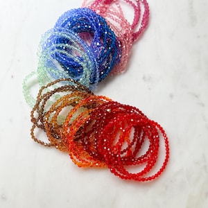 May include: A stack of colourful beaded bracelets in shades of red, orange, yellow, green, blue, and purple. The bracelets are made of small, faceted beads and are arranged in a rainbow pattern.