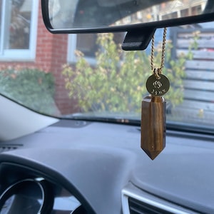 May include: A gold chain with a round charm featuring a paw print and the word "Lily" hangs from the rearview mirror of a car. A brown and gold tiger's eye crystal hangs from the charm.
