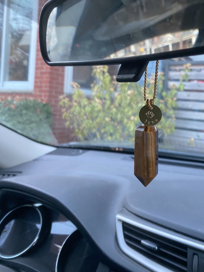 May include: A gold chain with a round charm featuring a paw print and the word "Lily" hangs from the rearview mirror of a car. A brown and gold tiger's eye crystal hangs from the charm.