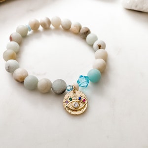 May include: A light blue and white beaded bracelet with a gold charm featuring an evil eye design with colorful gemstones.
