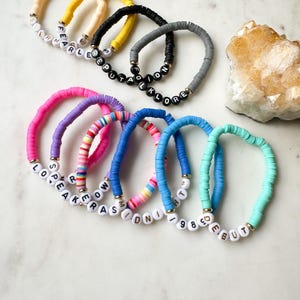 May include: A set of ten colorful bracelets with different words spelled out in white letters on black beads. The bracelets are made of colorful, round beads.