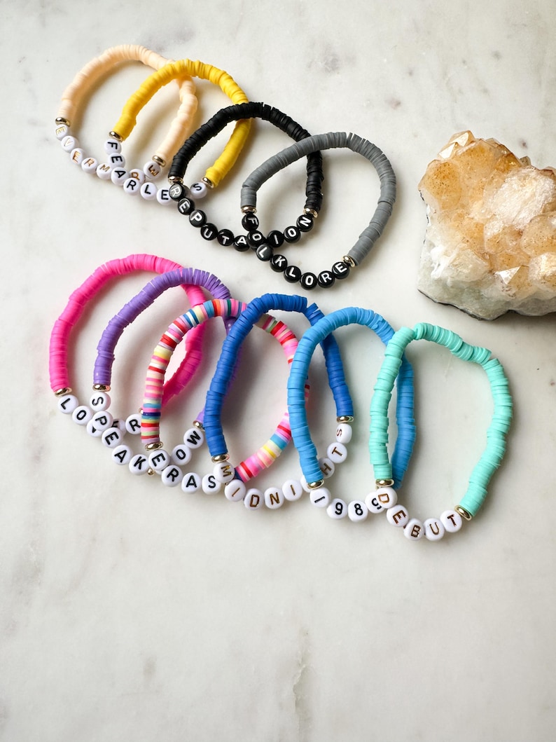 May include: A set of ten colorful bracelets with different words spelled out in white letters on black beads. The bracelets are made of colorful, round beads.