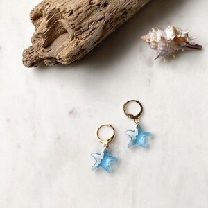 Puede incluir: Un par de pendientes de aro dorados con colgantes de estrella de mar azul claro. Los pendientes están sobre una superficie blanca.